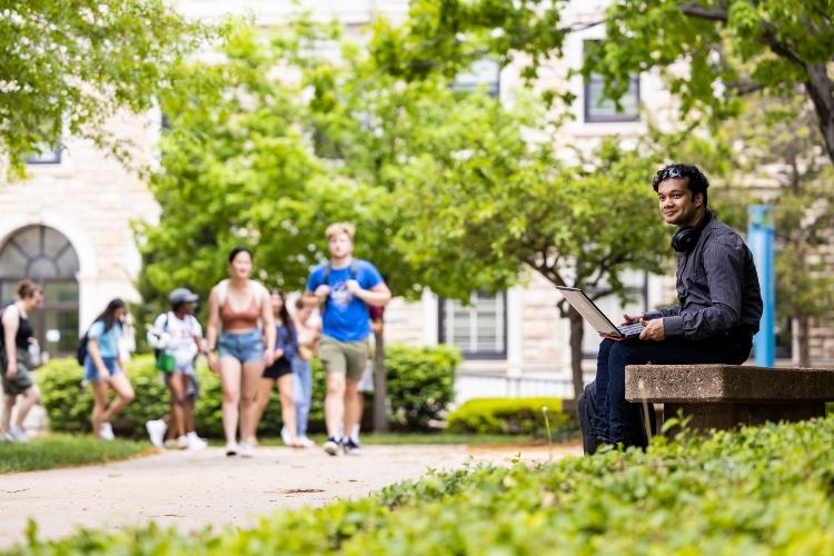 A student sits outside on a bench with a laptop on his lap. A group of students on the walkway behind him is walking his direction