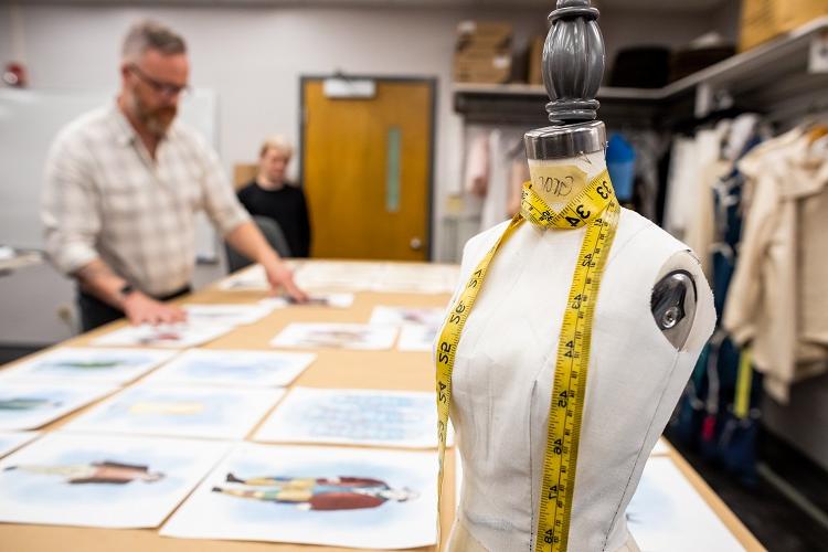 A mannequin with a measuring tape is in the forefront. Behind it is a large table with several ilustrations on it. A professor and student look at the illustrations.