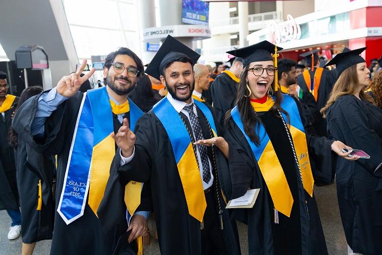 Three students smile exaggeratedly at the camera in full graduation regalia