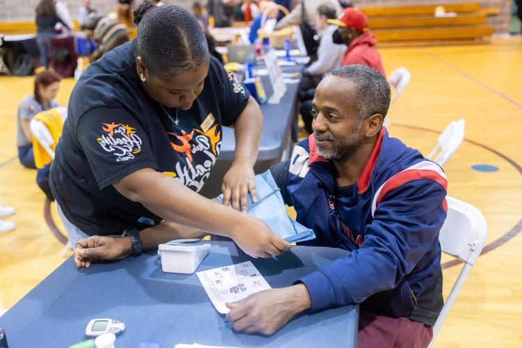 A person helps another person with a blood pressure cuff at a health event inside a gymnasium filled with people