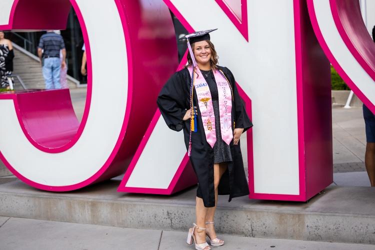 Macila Arnold stands in front of a large, bright pink KC sign smiling in a cap and gown