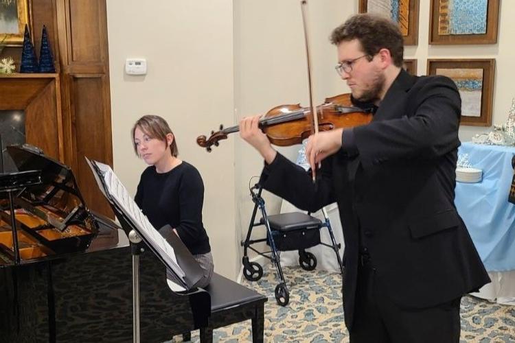 Beth plays a piano, a student plays the violin standing next to her