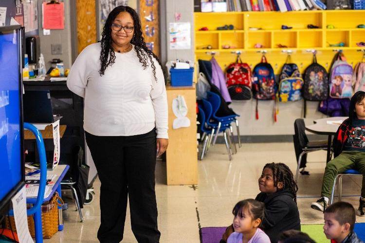 Kayla stands inside an elementary classroom with students looking up at a smartboard she's pointing to