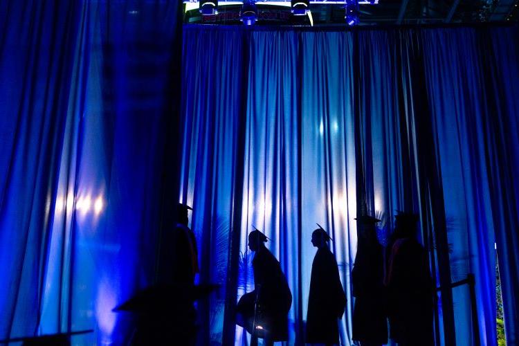 Graduates are silhouetted against blue curtains, waiting to walk across the stage